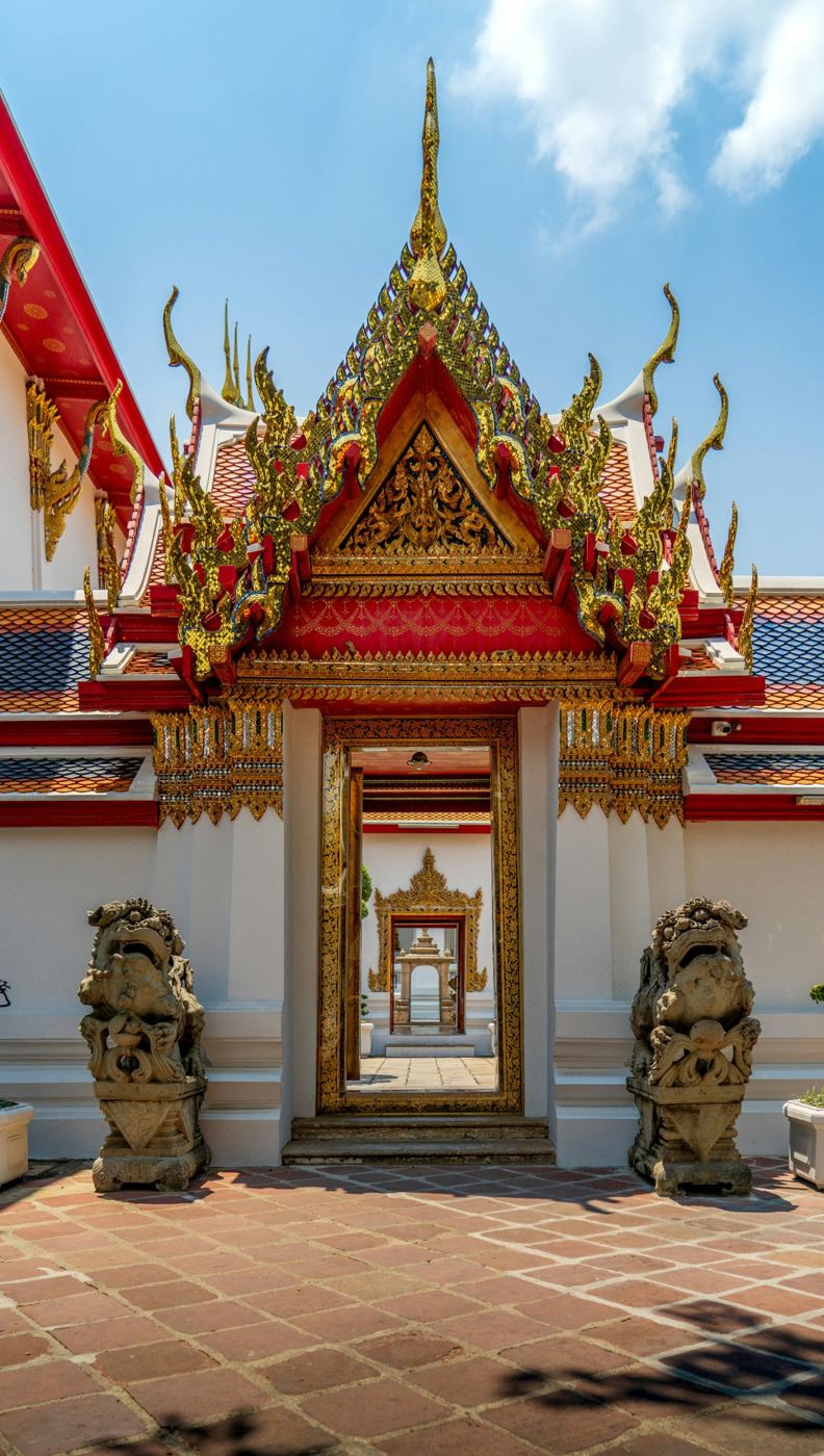 An ornate red and gold temple gateway guarded by two stone statues in Bangkok