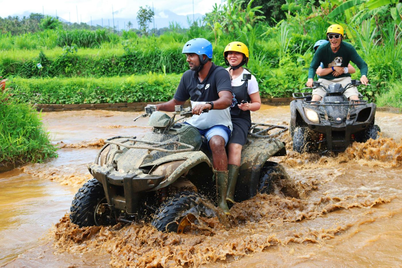 Riders on muddy ATVs splashing through a jungle stream