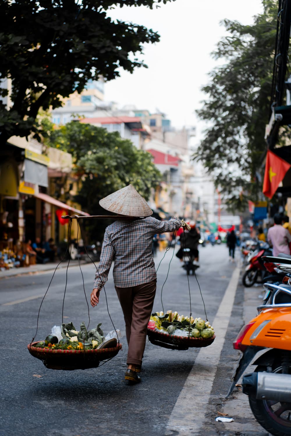 Street vendor in a conical hat carrying two baskets of vegetables down a Hanoi street
